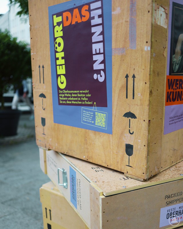 Eine weitere Seite der Kiste zeigt ein Plakat mit der Aufschrift „Gehört das Ihnen?“ vor städtischem Hintergrund.