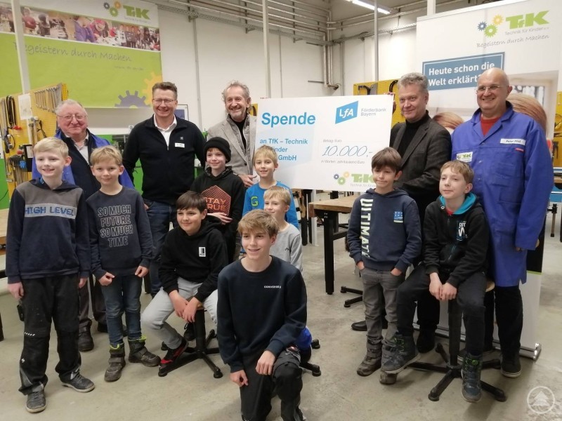 von links nach rechts: Alfred Tröger (Mentor TfK-Technikhaus Straubing), Werner Helmbrecht (Projektleiter TfK), Heinz Iglhaut (Vorstand TfK), Dr. Thomas Langer (Vorstandsmitglied LfA), Peter Pilsl (Mentor TfK-Technikhaus Straubing). Eine Gruppe aus Kindern und Erwachsenen posiert gemeinsam in einer Werkstatt. Mehrere Erwachsene halten einen großen Spendenscheck über 10.000 Euro.