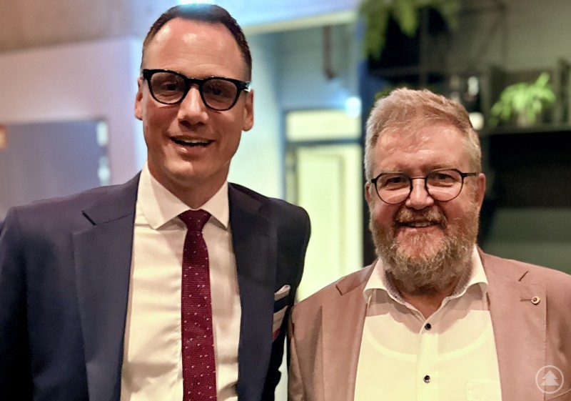 Prof. Dr. Christoph Juhn (v.l.) und Gerhard Drexler Zwei Männer in Anzug und Hemd stehen nebeneinander bei einem Netzwerkabend und lächeln in die Kamera, im Hintergrund moderne Innenarchitektur und Pflanzen.