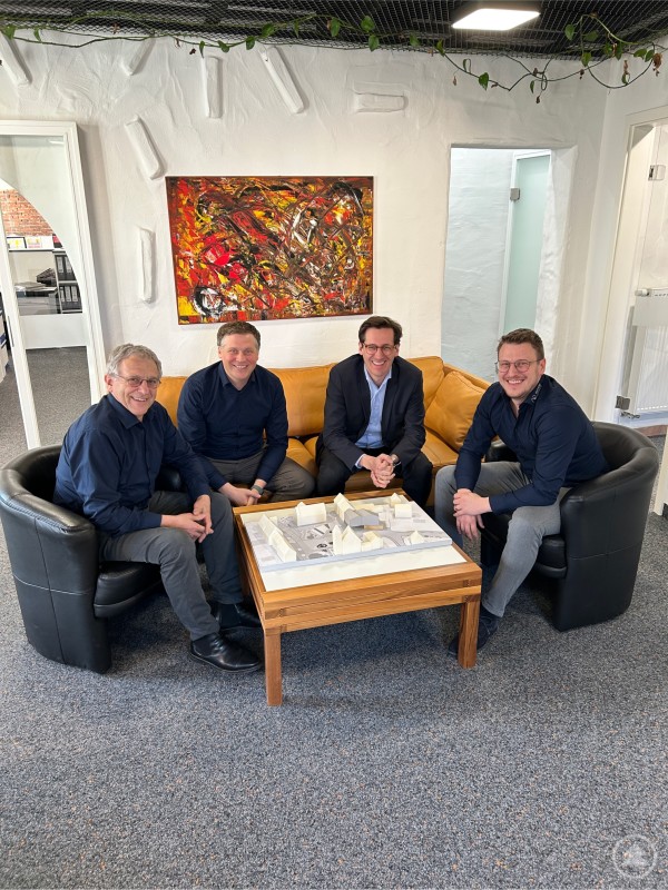 Landtagsabgeordneter Dr. Stefan Ebner (2.v.r.) informierte sich bei den Geschäftsführern Josef (l.), Johannes (2.v.l.) und Martin Weber (r.) von Weber Architekten über aktuelle kommunale Bauprojekte und die Herausforderungen des Planens und Bauens im ländlichen Raum. Vier Männer sitzen in einem Besprechungsraum rund um einen Tisch mit einem Architekturmodell. Im Hintergrund hängt ein großes farbenfrohes Kunstwerk an der Wand.