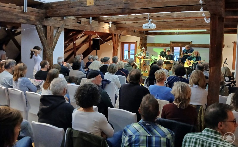 Die Band Missing-O-Love begeisterte die rund 90 Zuhörer mit feinem Folkrock der 60er- und 70er Jahre Das Bild zeigt ein stimmungsvolles Konzert in einem rustikalen Saal mit Holzbalken und gemütlichem Ambiente. Die Band „Missing-O-Love“ steht auf der kleinen Bühne und spielt vor rund 90 begeisterten Zuhörerinnen und Zuhörern. Mit mehreren Gitarren und Gesang bringen die Musiker feinfühligen Folkrock der 60er- und 70er-Jahre auf die Bühne. Das Publikum sitzt aufmerksam auf weißen Stühlen und genießt sichtlich die musikalische Atmosphäre. Die Mischung aus Live-Musik, Nostalgie und der besonderen Location macht den Abend zu einem besonderen Kulturerlebnis.