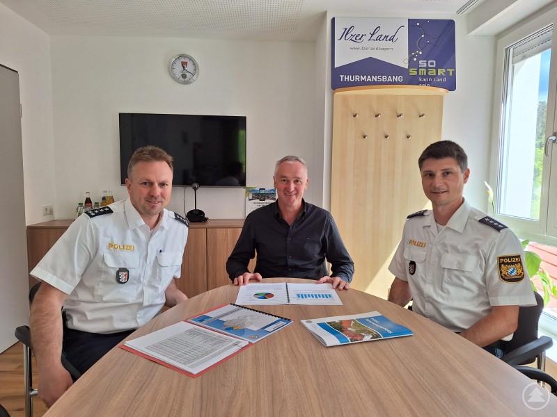 Drei Männer sitzen an einem Besprechungstisch in einem modern eingerichteten Büro. Zwei von ihnen tragen weiße Polizeiuniformen, der dritte in der Mitte ein dunkles Hemd. Vor ihnen liegen verschiedene Unterlagen mit Diagrammen und Statistiken.