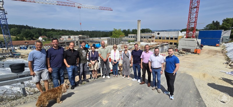ruppe von Politiker:innen und Projektverantwortlichen vor einer großen Baustelle mit Kränen und Baumaterialien; zwei Hunde sind ebenfalls zu sehen.