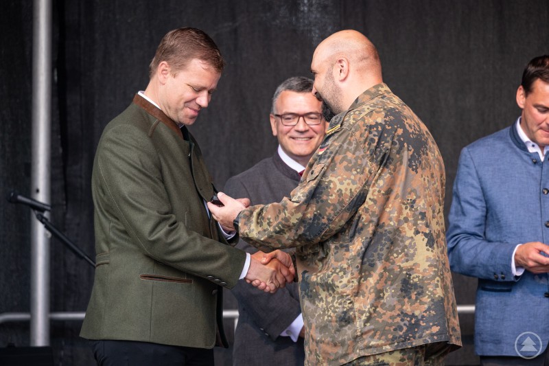 Ein Mann in Bundeswehr-Uniform überreicht einem anderen Mann im grünen Trachtenjanker eine Auszeichnung oder Medaille. Beide Männer reichen sich dabei die Hand. Im Hintergrund lächelt ein weiterer Mann in Tracht, rechts steht eine vierte Person in einem blauen Janker und schaut nach unten.