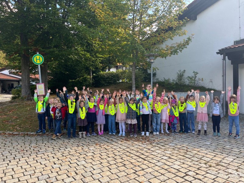 Eine große Gruppe von Grundschulkindern steht an einer Bushaltestelle, alle tragen leuchtend gelbe Sicherheitsüberwürfe und heben die Arme in die Höhe.