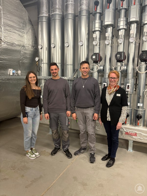 Vier Mitglieder des sechsköpfigen Energieteams der Kliniken Am Goldenen Steig (v.l.): Natalie Stiller (Marketing), Fabian Eibl (Assistent der technischen Leitung), Christian Siglmüller (Technische Leitung) und Anna Scherer (Qualitätsmanagement). Vier Mitarbeitende der Kliniken Am Goldenen Steig stehen in einem Technikraum vor mehreren großen, metallisch glänzenden Leitungen und Anlagen.