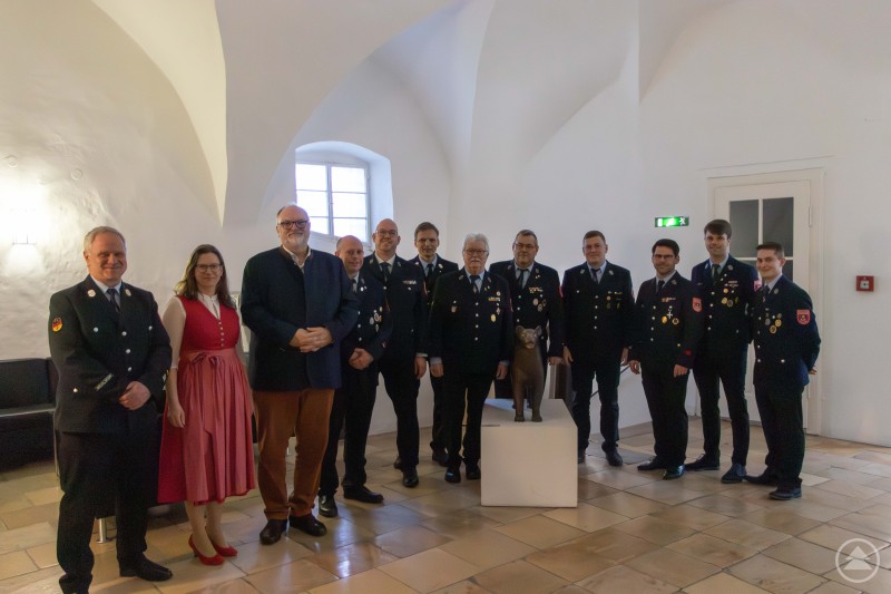 Drei M&auml;nner stehen in einem Raum vor einer Karte und einem Flipchart. In der Mitte h&auml;lt Oberb&uuml;rgermeister J&uuml;rgen Dupper eine Urkunde und ein Buchgeschenk in den H&auml;nden, w&auml;hrend zwei Vertreter der Freiwilligen Feuerwehr Grubweg in Uniform neben ihm stehen.