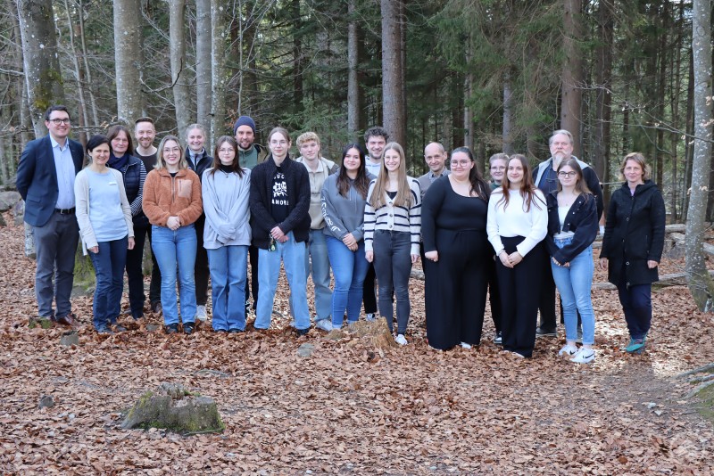 Nach der Projektvorstellung stellte sich die Klasse des Berufsbildungszentrums mit Schul- und Nationalparkvertretern zum Gruppenfoto auf. Das Bild zeigt eine gemischte Gruppe junger und älterer Personen bei einem Gruppenfoto im Wald. Sie stehen auf einer mit Laub bedeckten Lichtung zwischen hohen Bäumen und lächeln in die Kamera. Die Atmosphäre wirkt offen, freundlich und gemeinschaftlich. Es handelt sich vermutlich um ein Bildungs- oder Projektformat – passend zur Thematik „Leichte Sprache im Fokus“, etwa im Rahmen eines Workshops, eines inklusiven Projekts oder einer gemeinsamen Initiative von Bildungseinrichtungen und dem Nationalpark Bayerischer Wald. Die Verbindung von Natur, Bildung und gesellschaftlichem Engagement steht im Vordergrund.