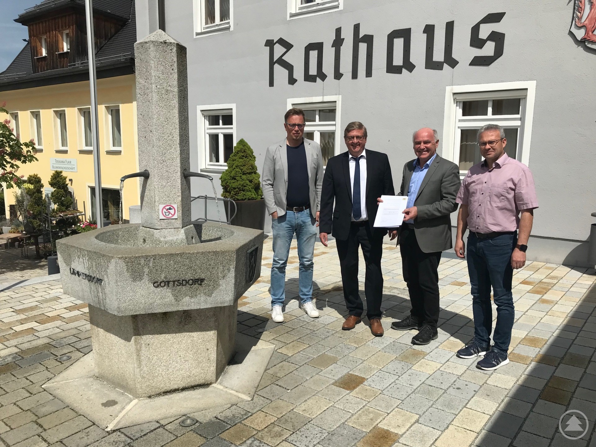 Vier Männer stehen vor dem Rathaus Untergriesbach neben einem Granitbrunnen, einer hält ein Dokument in der Hand.