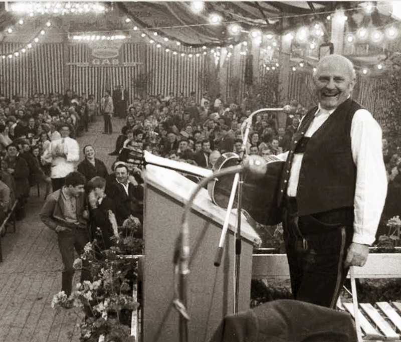 Kaum einer hatte die Menge so im Griff wie der Roider Jackl. Seine Gstanzl versetzten nicht nur die Festzelte bayernweit in Begeisterungsstürme. Ihm zu Ehren und um diese kulturelle Ausdrucksform in die Zukunft zu tragen, findet im Freilichtmuseum Massing ein Gstanzl-Kurs statt. Ein älterer Mann in traditioneller Tracht steht lachend an einem Rednerpult in einem voll besetzten Festzelt, das mit Lichterketten geschmückt ist.