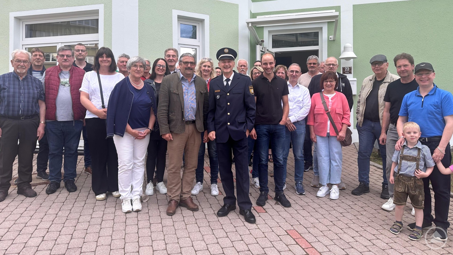 Eine Gruppe von rund 25 Personen, darunter Männer, Frauen und Kinder, steht vor einem hellgrünen Gebäude. In der Mitte trägt ein Mann eine Polizeiuniform.
