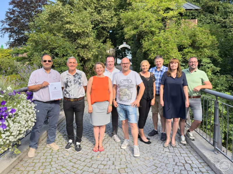 Die Vertreterinnen und Vertreter der Stadtgemeinde, des Tourismusvereins, von Schärding Innovativ und der Aktiwirte feiern die Gründung der Tourismus und Stadtmarketing Schärding GmbH. Eine Gruppe von neun Personen steht auf einem gepflasterten Weg vor einer grünen Parklandschaft. Eine Person hält ein offizielles Dokument in der Hand. Alle lächeln in die Kamera, es ist sonnig.