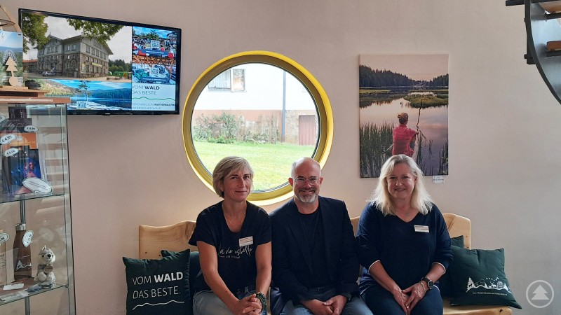 Robert Kürzinger (Mitte), Geschäftsführer der Ferienregion Nationalpark Bayerischer Wald, begrüßt die neuen Mitarbeiterinnen Kateřina Mosingerova (links) und Sabine Karl (rechts) im Team der Tourist Info Bayerisch Eisenstein. Zwei Frauen und ein Mann sitzen auf einer Holzbank vor einem runden Fenster, im Hintergrund touristische Werbemittel und Naturfotos – Innenraum einer Tourist Information.