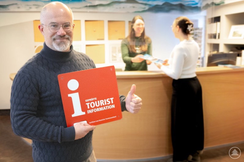 Robert Kürzinger, Geschäftsführer der Ferienregion Nationalpark Bayerischer Wald (FNBW), freut sich über die Auszeichnung mit der “i-Marke” und die damit verbundene Bestätigung der hohen Service- und Beratungsqualität aller zehn Touristinformationen der Ferienregion. Ein Mann mit Brille und grauem Rollkragenpullover steht in einer Touristinformation, hält ein rotes i-Marke-Schild hoch und zeigt mit dem Daumen nach oben. Im Hintergrund beraten zwei Mitarbeiterinnen eine Besucherin am Tresen.