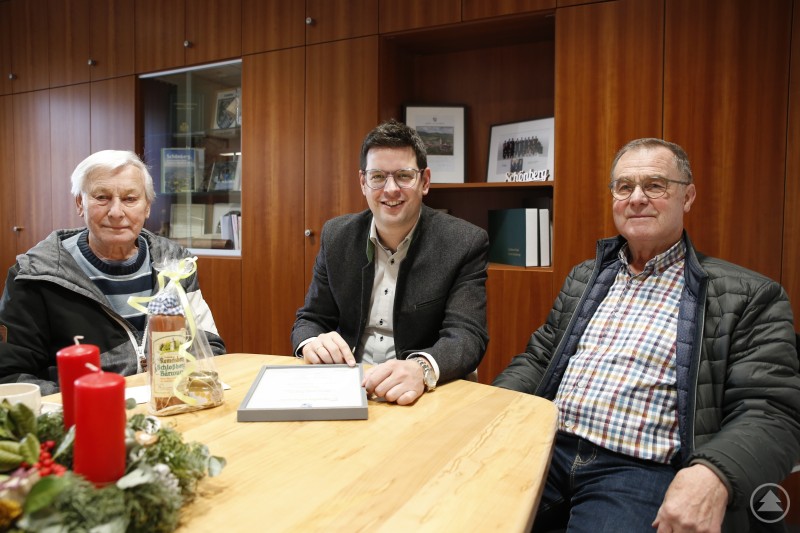 Ein herzlicher Dank für wissendes und zupackendes Ehrenamt ging an Josef Regener von Bürgermeister Martin Pichler und Obmann Johann Krammer. Drei Männer sitzen an einem Tisch in einem Besprechungsraum. In der Mitte liegt eine Urkunde, daneben stehen Kerzen und kleine Geschenke. Alle drei blicken in die Kamera.