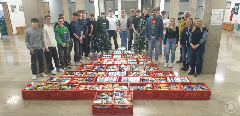 Stellvertretend für die Realschule Grafenau übergab die Klasse 10a über eine Tonne Lebensmittel an die Grafenauer Tafel (v. rechts Suzanne Mayer von der Tafel Grafenau, Religionslehrkraft und Organisatorin Stephanie Müller-Peter, Hausmeister Gerhard Siegl, Schulleiter Ferdinand Klingelhöfer) Eine Gruppe von Schülerinnen und Schülern der Staatlichen Realschule Grafenau steht gemeinsam mit Lehrkräften und Vertretern der Grafenauer Tafel in einer Aula. Vor ihnen sind zahlreiche rote Kisten mit haltbaren Lebensmitteln in Kreuzform aufgebaut, im Hintergrund stehen zwei geschmückte Weihnachtsbäume.