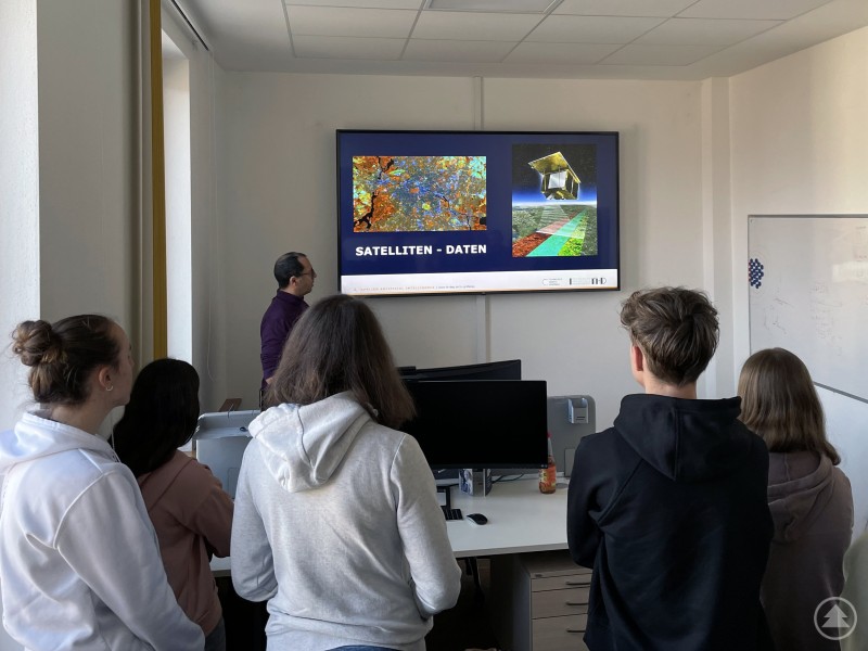 Michael Fernandes, wissenschaftlicher Mitarbeiter am TCG, zeigt den Schülerinnen und Schülern, wie der Weltraum von Grafenau aus erforscht wird. Das Bild zeigt eine Gruppe Jugendlicher bei einer Präsentation im Rahmen der Wirtschaftswoche am Landgraf-Leuchtenberg-Gymnasium (LLG). Die Schüler*innen stehen in einem modernen Klassen- oder Technikraum vor einem Bildschirm, auf dem eine Folie mit dem Titel „SATELLITEN – DATEN“ zu sehen ist. Die Präsentation thematisiert offensichtlich Satellitenbilder und deren Auswertung, vermutlich im Kontext von Digitalisierung, Umweltbeobachtung oder künstlicher Intelligenz. Der Vortragende erklärt anschaulich die gezeigten Inhalte. Die Szene vermittelt Interesse an Zukunftsthemen und praxisnaher Bildung in einem innovativen schulischen Umfeld.
