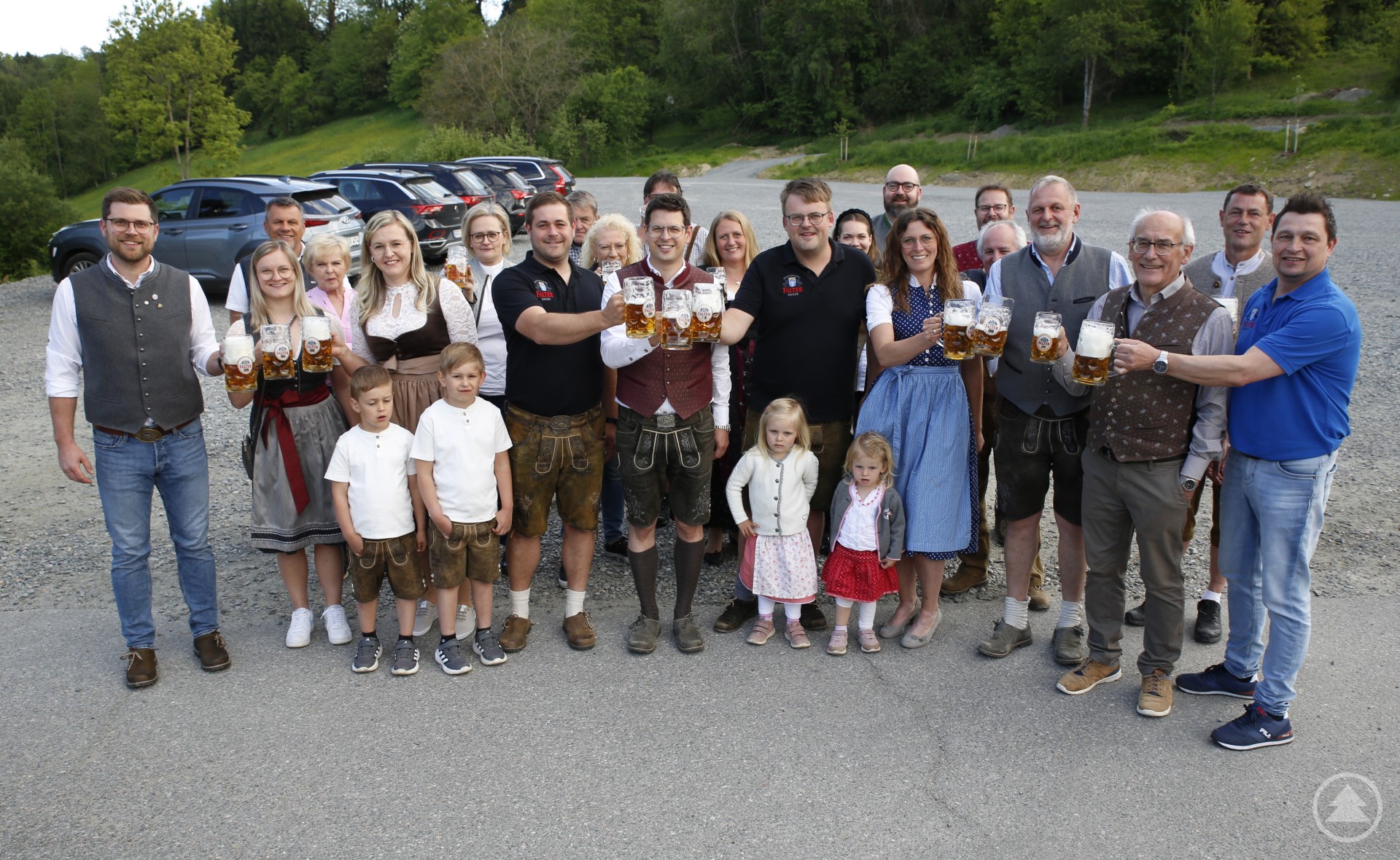 Vertreter der Brauerei und der Festwirte stoßen gemeinsam auf das erfolgreiche Falter-Festbier an, das beim Test die Erwartungen voll erfüllt hat. Eine Gruppe von Menschen, darunter Festwirte, Brauereivertreter und Gemeinderäte, stoßen mit Maßkrügen auf die erfolgreiche Bierprobe des Falter-Festbiers an. Im Hintergrund ist eine grüne Wiese und ein Parkplatz zu sehen.