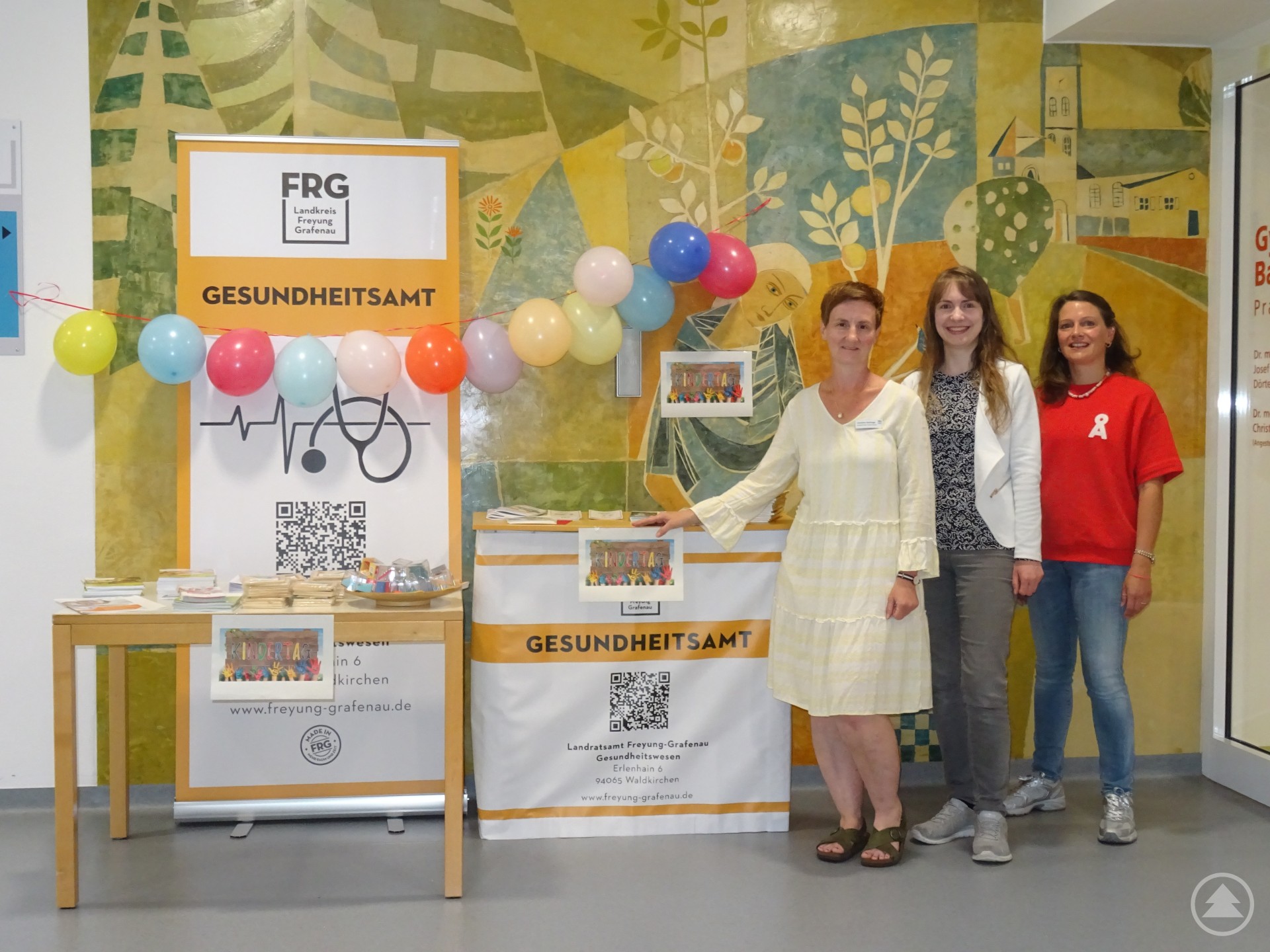 Drei Frauen stehen neben einem bunt dekorierten Infostand des Gesundheitsamts im Eingangsbereich eines Gebäudes. Auf dem Tisch liegen Infomaterialien, im Hintergrund hängen Luftballons.