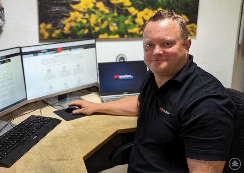 Stefan Behringer, Geschäftsführer der BWmedien GmbH, arbeitet im Büro an der Weiterentwicklung von BWverein – der digitalen Plattform für moderne Vereinsverwaltung. Stefan Behringer sitzt an einem Schreibtisch mit drei Bildschirmen und blickt in die Kamera. Auf den Monitoren ist die Verwaltungssoftware BWverein zu sehen, im Hintergrund ein Wandbild mit gelben Blumen.