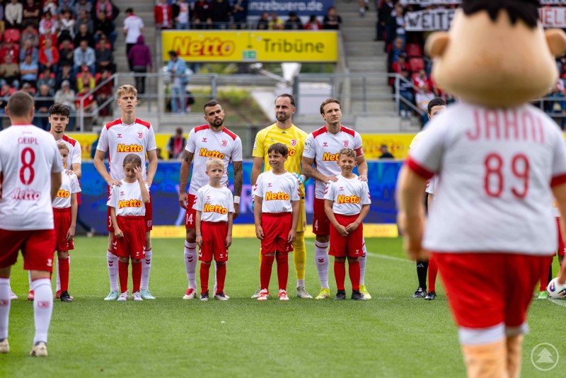Das Bild zeigt Fußballspieler in weißen Trikots mit rotem Aufdruck und roten Hosen, die gemeinsam mit Kindern in ähnlichen Outfits auf dem Spielfeld stehen. Im Vordergrund ist ein Maskottchen mit der Rückennummer 89 und dem Namen „JAHN“ zu sehen. Im Hintergrund sitzen Zuschauer auf der Tribüne, über der große gelbe Werbebanner mit der Aufschrift „Netto“ hängen. Es wirkt wie die Aufstellung der Mannschaft vor dem Anpfiff eines Spiels.