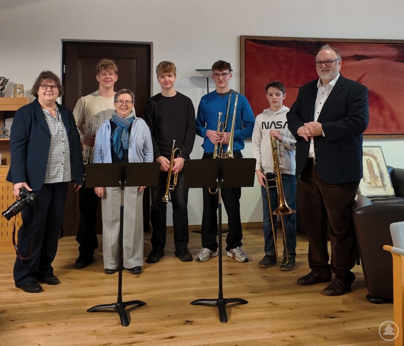 Oberbürgermeister Jürgen Dupper (rechts) dankt den Schülern sowie den Lehrkräften für den Besuch und die dargebotene Weihnachtsmusik. Mehrere Schülerinnen und Schüler des Auersperg-Gymnasiums stehen gemeinsam mit ihren Lehrkräften und Oberbürgermeister Jürgen Dupper in einem Raum. Die Jugendlichen halten Trompeten und Posaunen in den Händen, vor ihnen stehen Notenständer. Die Gruppe posiert nach dem musikalischen Vortrag in ruhiger, festlicher Atmosphäre.