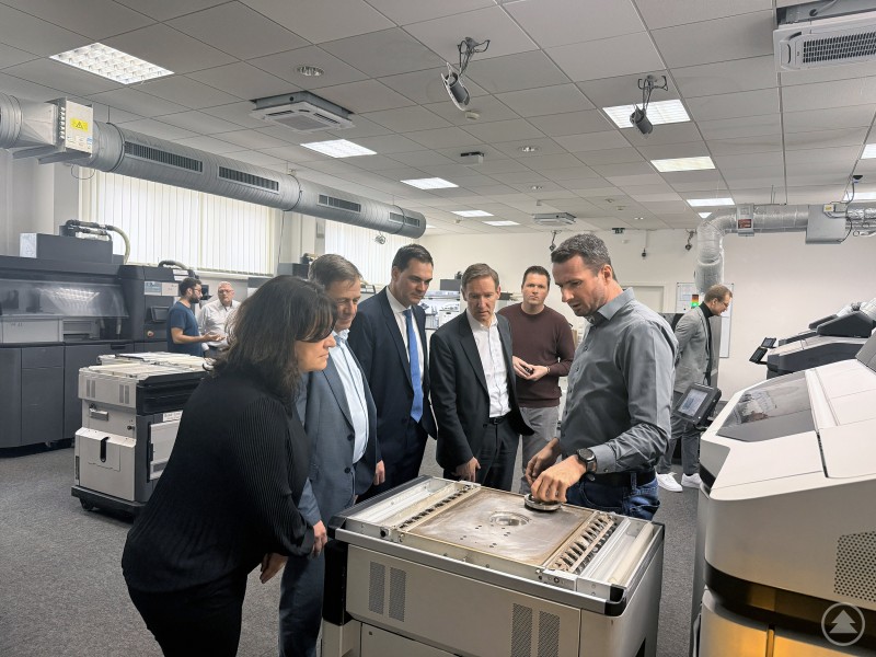 Eine Gruppe von sechs Personen, &uuml;berwiegend in Anzug, beobachtet die Vorf&uuml;hrung einer industriellen Maschine in einer Werkhalle. Im Hintergrund sind weitere Personen und moderne Produktionsmaschinen zu sehen.