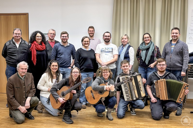Die Vertreter der teilnehmenden Institutionen von Bezirk Niederbayern, Bayerischem Landesverein für Heimatpflege e.V., Kreisvolksmusikpflege der niederbayerischen Landkreise, Volksmusikakademie Bayern und Trachtengau Niederbayern tauschten sich mit Schülerinnen und Schülern des Ausbildungszweigs Volksmusik aus. Gruppenfoto in einem Saal: Vertreterinnen und Vertreter verschiedener Volksmusik-Institutionen stehen und sitzen mit Schülerinnen und Schülern, vorne sind Gitarren und ein Akkordeon zu sehen.