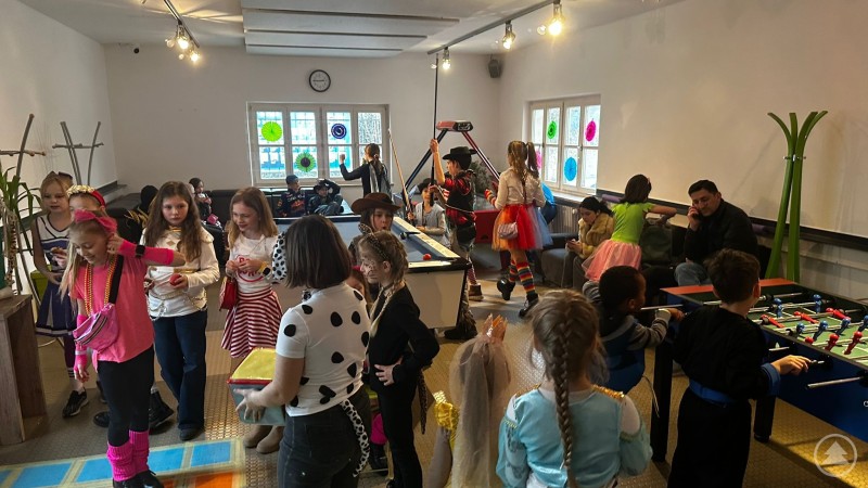 Zahlreiche Kinder in bunten Faschingskost&uuml;men spielen und tanzen im Jugendcenter 4You, einige stehen am Billardtisch und Tischkicker, andere unterhalten sich oder probieren Spiele aus.