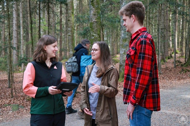 Bei der letzten Besucherbefragung zeigten sich 85 Prozent der Nationalparkbesucher sehr zufrieden. Das Bild zeigt drei junge Erwachsene im Gespräch auf einem Waldweg – vermutlich im Rahmen einer Besucherumfrage oder einer naturpädagogischen Aktion. Eine der Personen hält ein Tablet, was auf eine digitale Erhebung oder Datenerfassung schließen lässt. Alle wirken freundlich und interessiert. Im Hintergrund spaziert eine weitere Person mit Rucksack durch den Wald. Die Szene vermittelt eine offene, moderne Form der Umweltbildung oder Besucherbetreuung in einem Naturpark oder Schutzgebiet.