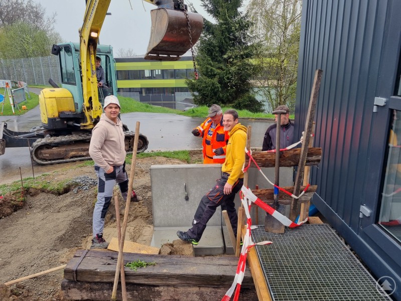 Mit LEADER-Förderung und ehrenamtlichem Einsatz wurde 2023 die Außenanlage am Vereinsheim des TV Freyung neugestaltet. Mehrere Männer arbeiten bei Nieselregen auf einer Baustelle an einem Gebäude mit dunkler Fassade. Ein Bagger hebt ein Betonteil an, Werkzeuge und Absperrbänder sind sichtbar.