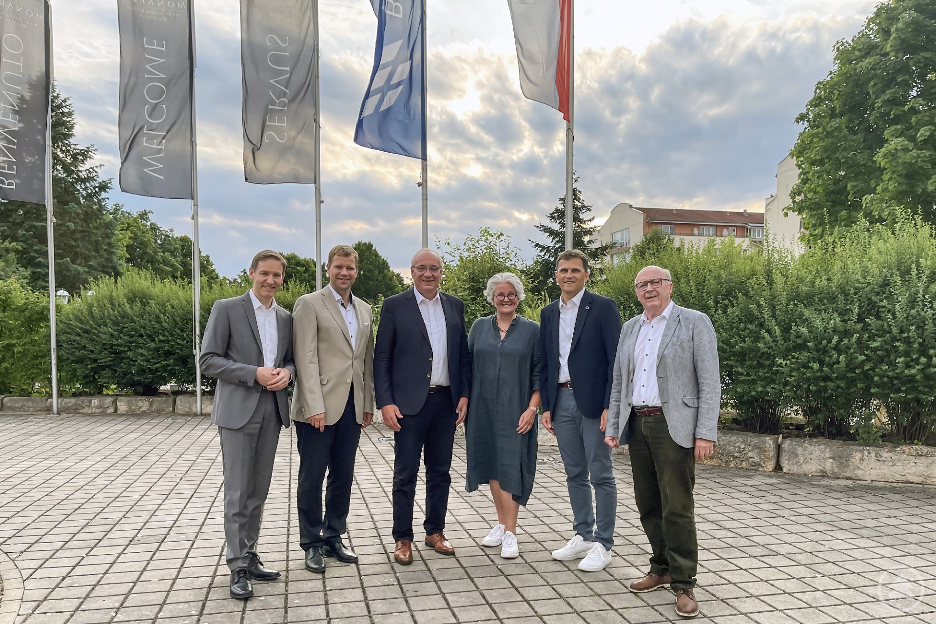 Sechs Personen stehen in einer Reihe im Freien vor mehreren Fahnenmasten. Es handelt sich um Vertreter aus Politik und Verwaltung, die an der Vollversammlung des Bayerischen Bezirketags teilnehmen. Im Hintergrund sind grüne Büsche und ein teilweise bewölkter Himmel zu sehen.