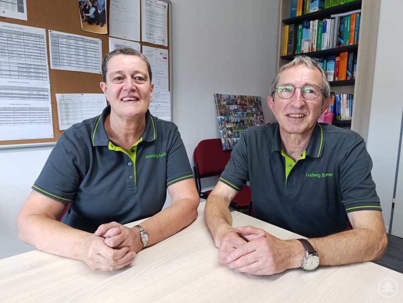 Astrid Schricker und Ludwig Brunner sitzen gemeinsam im Büro und wirken gut gelaunt – ein eingespieltes Team, das Kompetenz und Freundlichkeit ausstrahlt. Zwei Personen sitzen an einem Tisch in einem Büro und lächeln in die Kamera.