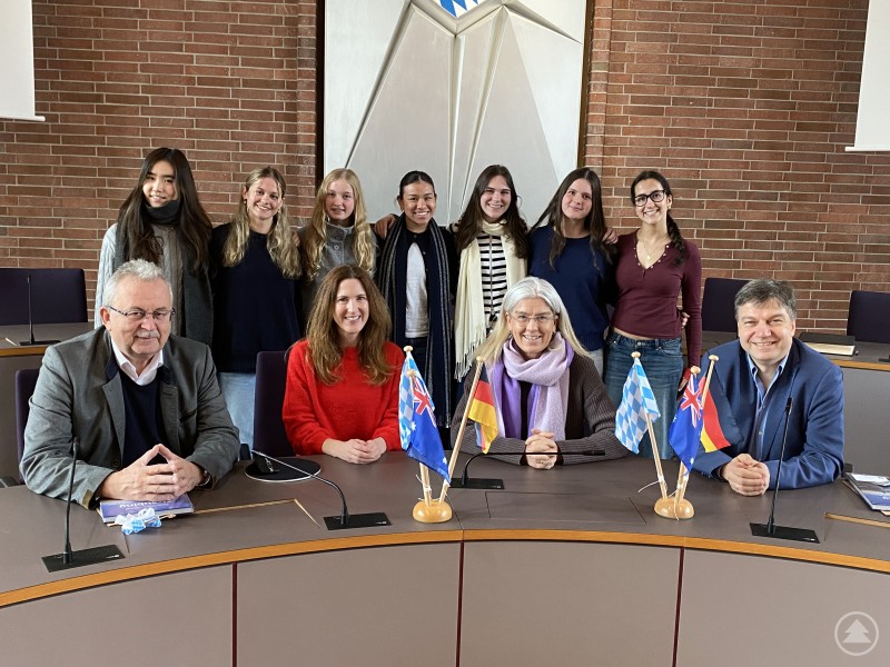 Die australischen Gastschülerinnen im Sitzungssaal des Landratsamts mit ihren Begleitlehrerinnen, Simon Wech und Landrat Josef Laumer. Eine Gruppe australischer Gastschülerinnen steht im Sitzungssaal des Landratsamtes hinter einem Tisch, an dem vier Erwachsene sitzen. Auf dem Tisch stehen mehrere kleine Länderfähnchen. Alle Personen blicken freundlich in die Kamera.