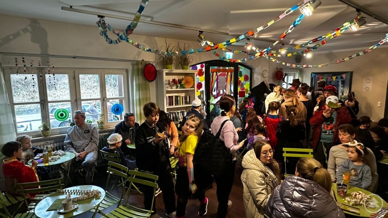 Gut besuchter Veranstaltungsraum im Jugendcenter 4You mit verkleideten Kindern und Eltern, bunte Girlanden schmücken die Decke, an Tischen wird gegessen und gelacht.