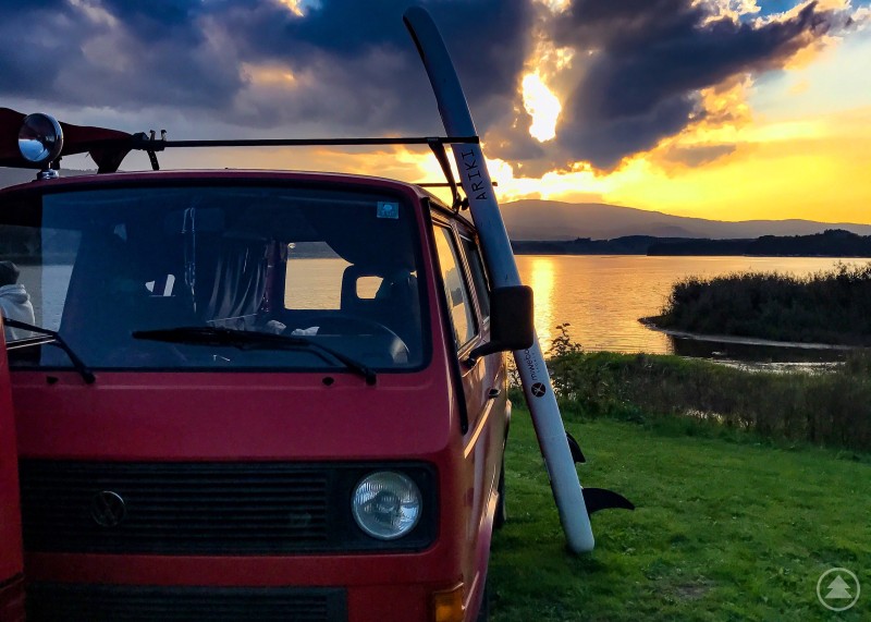 Farbgewaltiges Sonnenuntergangspanorama an einem See mit VW Bus und Paddleboard im Vordergrund.