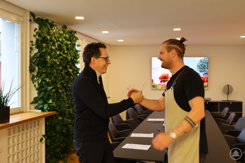 Zwei M&auml;nner stehen sich in einem Besprechungsraum gegen&uuml;ber, l&auml;cheln und geben sich die Hand. Im Hintergrund sind Pflanzen und ein gro&szlig;er Tisch zu sehen.