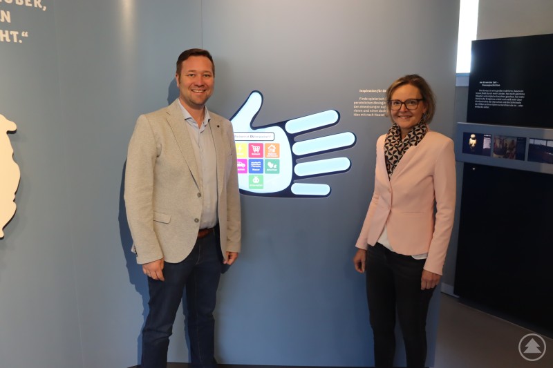 Der Geschäftsführer der Umweltbildungseinrichtung „Haus am Strom“, Josef Heisl (MdL), begrüßt die Caritasvorständin Andrea Anderlik. Die Caritas ist ist jetzt Gesellschafterin. Das Bild zeigt zwei Personen, einen Mann und eine Frau, die in einer Ausstellung oder einem modernen Informationsraum vor einer interaktiven Wand stehen. Die Wand ist in Form einer stilisierten Hand gestaltet, in deren Mitte bunte Symbole zu Themen wie Energie, Konsum, Mobilität, Wasser, Ernährung und Mitmachen zu sehen sind – vermutlich in Bezug auf Nachhaltigkeit oder Umweltschutz. Beide Personen lächeln in die Kamera. Die Szenerie wirkt offiziell und gleichzeitig einladend – wahrscheinlich im Rahmen einer Eröffnung, eines Besuchs oder einer Präsentation zu einem Bildungs- oder Umweltprojekt.