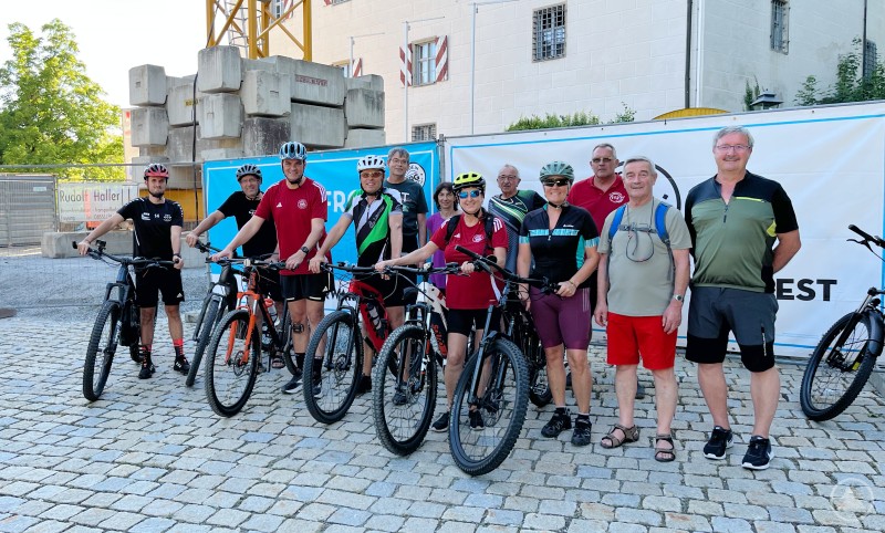 Rund ein Dutzend Radlbegeisterte machten sich bei rund 30 Grad auf eine gemeinsame Tour mit Landrat Sebastian Gruber (3. v. l.) und Koordinator Johannes Weiner (ganz links). Eine Gruppe von Radfahrerinnen und Radfahrern mit ihren Rädern vor einer Baustelle und einem Schloss, Landrat Gruber in rotem Trikot mittig.