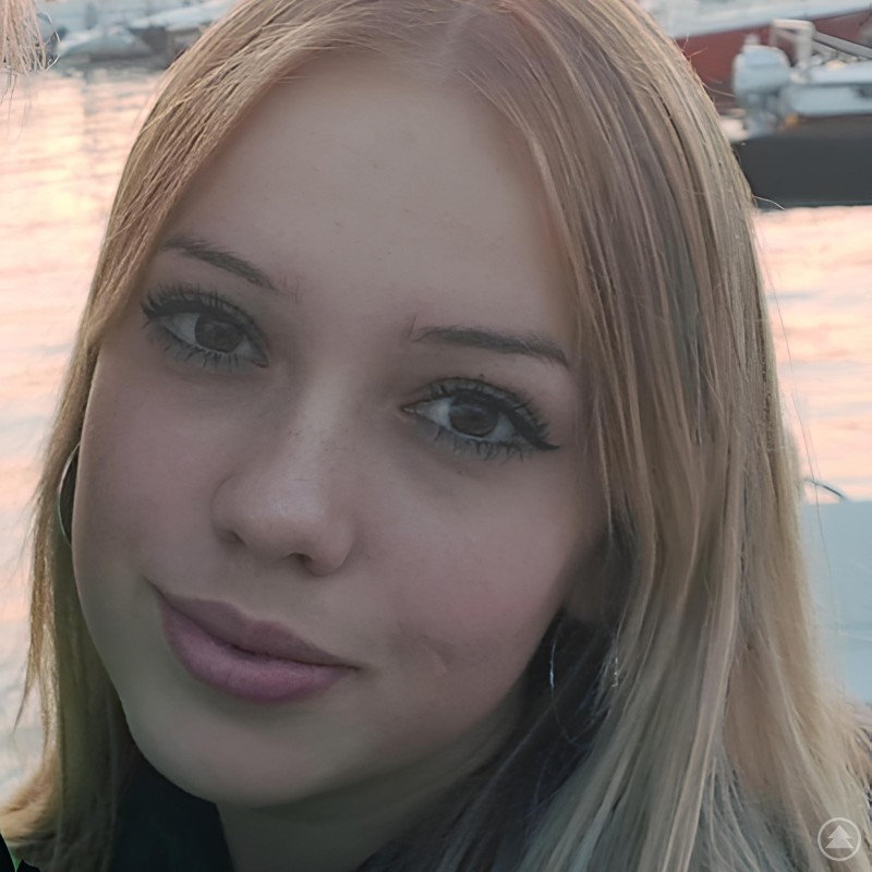 Junge Frau mit blonden Haaren blickt in die Kamera, im Hintergrund ein Hafen mit Booten bei Sonnenuntergang.