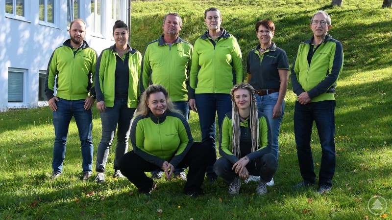 Acht Personen in grüner und grauer Kleidung stehen und knien auf einer Wiese vor einem Gebäude.