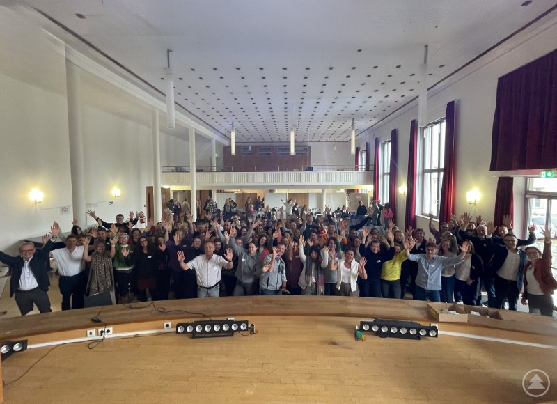 Herzliches Willkommen bei der Caritas: Rund 120 neue Mitarbeiter:innen starteten mit einem Einführungstag in ihre sinnstiftende Tätigkeit. Ein großer Saal voller Menschen, die fröhlich in die Kamera winken. Die Teilnehmenden stehen dicht gedrängt und strecken die Hände nach oben – eine fröhliche, gemeinschaftliche Atmosphäre.