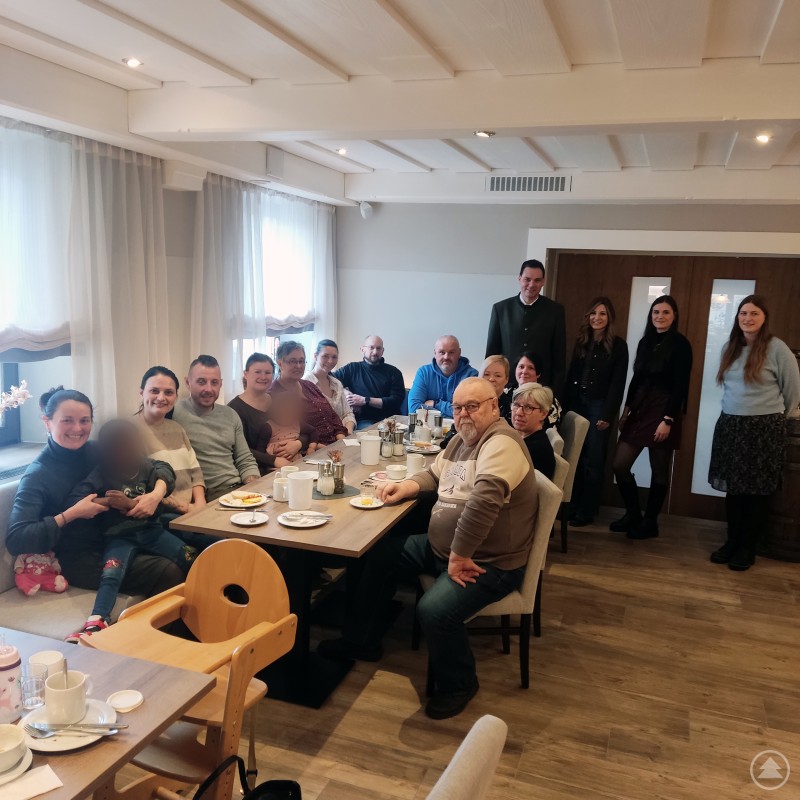 Engagierte Pflegeeltern aus dem Landkreis tauschten sich beim Stammtisch in Freyung aus. Landrat Sebastian Gruber dankte für ihren wichtigen Einsatz. Mehrere Pflegeeltern sitzen gemeinsam an einem Tisch in einem Gasthaus und frühstücken, im Hintergrund stehen Landrat Sebastian Gruber und weitere Beteiligte für ein Gruppenfoto.