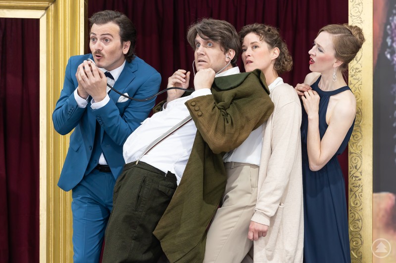 Vier Schauspieler in ausdrucksstarker Pose, einer hält ein Stethoskop, die Szene spielt auf einer farbenfrohen Theaterbühne.