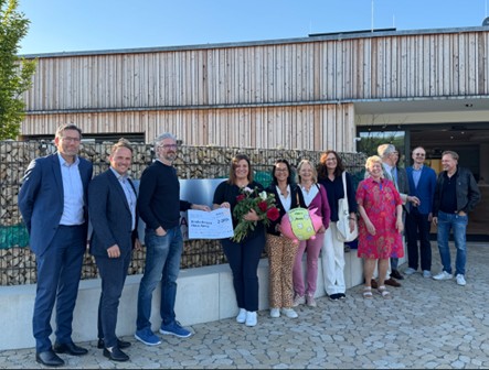 Eine Gruppe von rund einem Dutzend Personen steht vor einem modernen Gebäude aus Holz und Stein. In der Mitte wird ein großer Spendenscheck überreicht, eine Frau hält zudem einen Blumenstrauß und ein Plüschtier mit Herzsymbol.