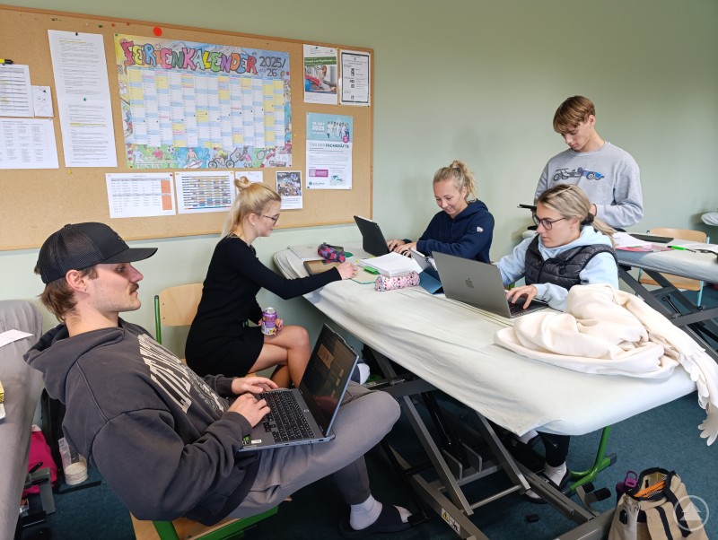 Im Unterricht herrscht konzentrierte, aber lockere Stimmung – die Schülerinnen und Schüler arbeiten gemeinsam an ihren Aufgaben und nutzen moderne Technik. Mehrere Schüler sitzen und stehen an Behandlungsliegen und arbeiten an Laptops und Notizen.