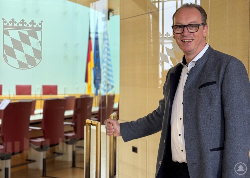 MdL Martin Behringer im Eingangsbereich des Plenarsaals, MdL Martin Behringer steht lächelnd im Eingangsbereich eines modernen Sitzungssaals. Hinter ihm sind ein großer Tisch, Stühle und bayerische Flaggen zu sehen.