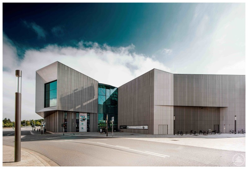 Ein modernes, architektonisch auffälliges Gebäude mit klaren Linien und großen Glasflächen. Die Südansicht zeigt den Haupteingang des Museums mit Plakaten und Fahrradständern – das Ensemble wirkt einladend und futuristisch.