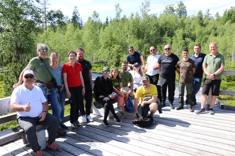 Die rumänische Delegation rund um Christoph und Barbara Promberger informierte sich gemeinsam mit Nationalparkleiterin Ursula Schuster und weiteren Gästen am Naturerlebnis Wistlberg über den Nationalpark Bayerischer Wald. Gruppe von rund 16 Personen auf einem Holzsteg im Grünen, bei sonnigem Wetter, lächelnd in die Kamera blickend.