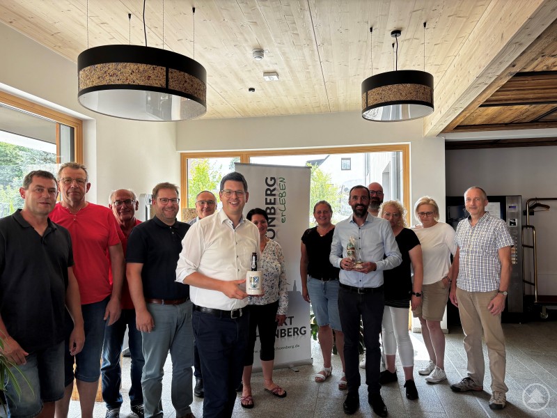 Reger Austausch zwischen den Mitgliedern des Marktgemeinderats Schönberg und den Gastgebern in Moosbach bei der diesjährigen Klausur. Eine Gruppe von etwa 14 Personen steht in einem modernen Raum mit Holzdecke. Zwei Männer in der Mitte halten Geschenke in den Händen. Im Hintergrund ist ein Roll-Up mit dem Schriftzug „Schönberg erleben“ zu sehen.